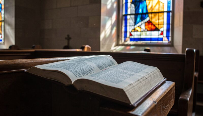 An open hymnal resting on a wooden pew with soft morning light falling through a blue and amber stained glass window.